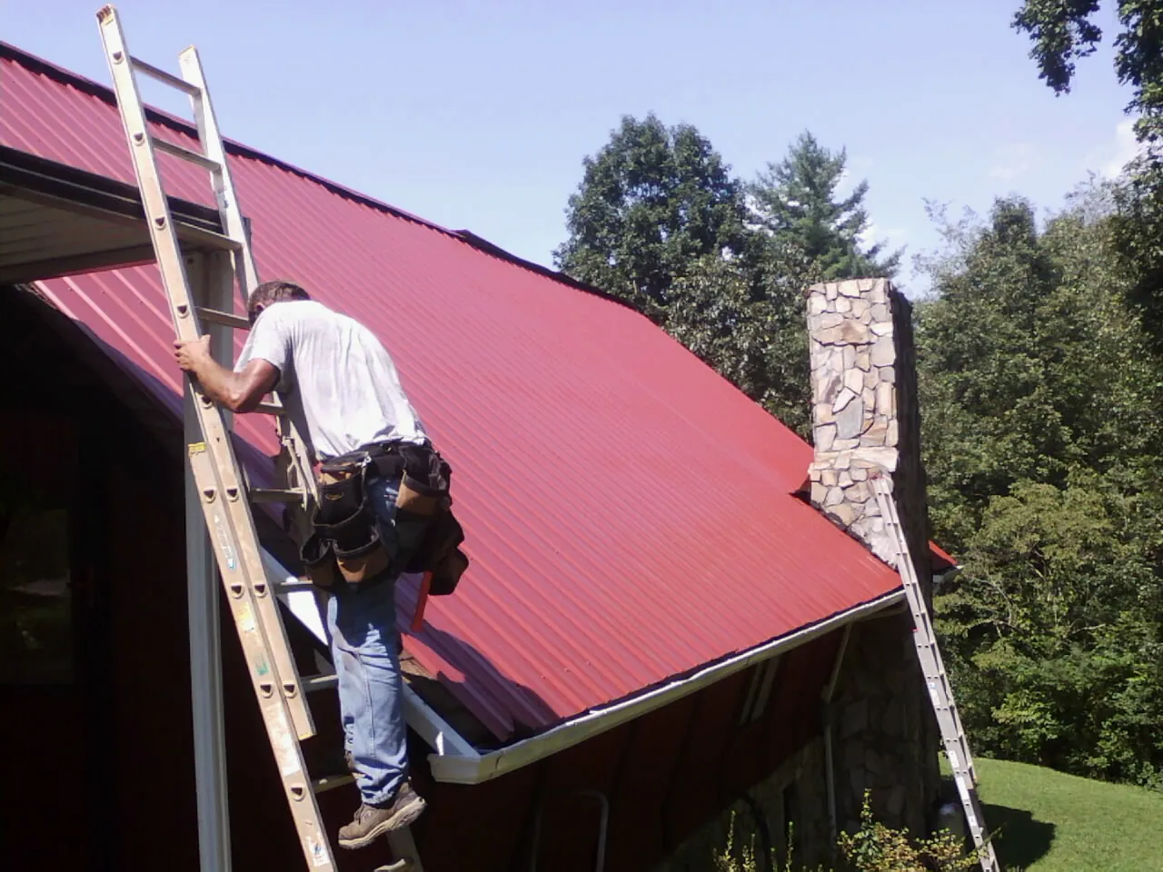 Roofer on a ladder beside a red metal roof and stone chimney — Soffit & Fascia Repair by Roofing Brothers in Bergholz